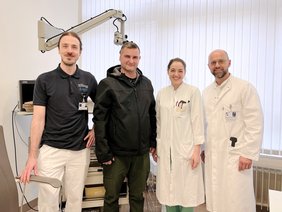 Im Behandlungszimmer, Patient mti schwarzer Kapuzenjacke, Seitenscheitel. Links Audiologie mit blauem Poloshirt, Zopf mit Mittelscheitel, etwas Bart, Grübchen, die Ärzten mit braunen zurückgebundenen Haaren. kleinen Kreolen, Arztkittel, der Arzt mit 3-Tagebart, Brille, Arztkittel ohne Kopfhaare, alle lächelnd