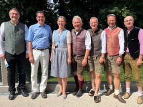 Mitanand 2025 (© Veronika Biller) Vier Männer rechts mit Lederhose, Hemd, Weste und Haferlschuhe, daneben die Dame in blauem Dirndl, die zwei Herren links mit bayerischer Weste (Prof. Weigert) und Prof. Rust mit blauem Hemd und weißer Hose. Alle im Garten mit Sonnenschein und lachend