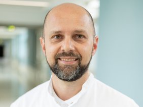 Portrait eines Mannes mit wenig Oberhaar und Vollbart ohne Brille und Kittel, lachend mit hellblauem Krankenhausflur im Hintergrund