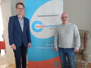 Rezertifizierung Wundexperte (© Sabine Muck) Zwei Herren vor einem blau-weißen Roll Up, Dr. Giesler im blauen Anzug, Herr Siebert mit Jeans und grauen Pulli