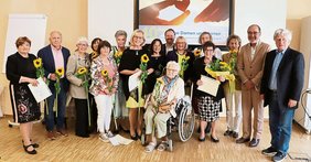 40 Jahre Gelbe Damen und Herren (© Monika Schneider-Stranninger) Alle Gelben Damen und Herren mit Sonnenblume in der Hand, eine Dame in Rollstuhl vorne, Frau Messerschmitt mit Blumenstrauß. Manche mit ihren überreichten Urkunden in der Hand oder am Boden. Sie stehen vor einem großen Bildschirm.