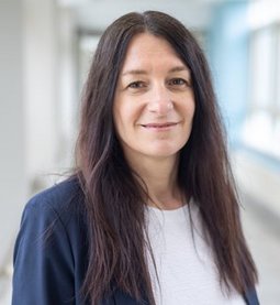 Dame mit langen braunen Haaren, dunkelblauem Blazer und weißem Shirt, lächelnd