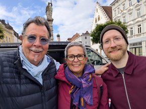 Hörbus Straubing 2025 (© Klinikum Straubing) Selfie auf dem Stadtplatz, mit Brunnen und blauem Himmel im Hintergrund. Herr Roth mit Sonnenbrille, blauer Steppjacke und hellblauem Schal, grauem Schnurrbart und ebensolche Haare. Die Damen mit lila Schal und violetter Jacke, kurze graue Haare und Brille, rechts mit Mütze, Brille und rotem Vollbart sowie roter Zipweste