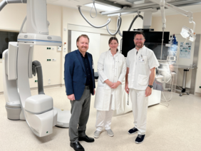 Überregionale Stroke Unit - TEMPiS Netzwerk (© Sandra Wimmer) Der Geschäftsführer in dunkelblauem Sacko, schwarzem Hemd und Schuhen und grauer Hose neben den beiden Ärzten in weiß vor der futuristisch anmutenden Anlage