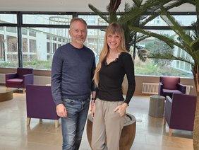 Ein Herr mit kurzem Vollbart, Jeans und blauem Pullover links mit einer jungen Frau mit blonden langen Haaren, beiger Hose und schwarzem Longsleeve vor lila Sessel und Palme im Foyer des Klinikums