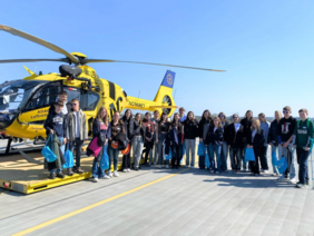 Tag der Ausbildung Landeplatz (© Marion Sieber) Etwa 25 Schüler in einem Alter von etwa 15-16 Jahren vor dem gelben ADAC-Hubschrauber am Landeplatz mit blauen Stoffrucksäcken, rechts der Junge mit Krücken. Blauer Himmel, Sonnenschein, der die Schülerinnen und Schüler blendete.