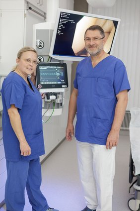 Endoskopie (© Johannes Lehner) Frau Probst ganz in blauer Arbeitskleidung, blond, mit Brille und Pferdeschwanz, kleiner als Prof. Dr. Weigert in weißer Hose und blauem Oberteil, vor zwei Bildschirmen