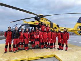Etwa 15 Männer und Frauen in roter Luftretteruniform vor dem gelbem ADAC-Hubschrauber am Landeplatz auf dem Dach des Klinikums. Ein Kollege hängt für das Foto an der Rettungswinde. Alle lächeln.
