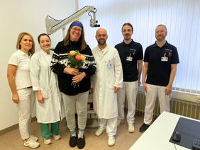 Die Patientin mit blauem Verband,wie ein Stirnband getragen, mit Brille, Hausschue, mittellange Haare, dunkler Norwegerpulli und Hausschuhe einen Strauß haltend, lacht. die junge Ärztin und der Chefarzt mit Vollbart und Brille in Kittel, Frau Gutjar auch in weiß, die Audiologen in dunkelblauem Polo. 
