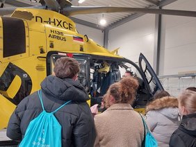Schüler von hinten mit türkisen Stoffrucksäcken mit Klinikumslogo, die vor dem gelben ADAC-Hubschrauber stehen, dessen Tür geöffnet ist. Ein Notarzt mit Brille steht davor.