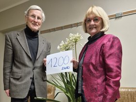 (©Förderverein) Zwei Personen, eine Frau und ein Mann, stehen nebeneinander in einem Innenraum. Beide lächeln in die Kamera und halten gemeinsam ein weißes Blatt Papier mit der handschriftlichen Aufschrift „1200 Euro“. Zwischen ihnen steht eine grüne Zimmerpflanze in einem Topf auf einem kleinen Tisch. Die Szene vermittelt eine Spendenübergabe in freundlicher Atmosphäre.