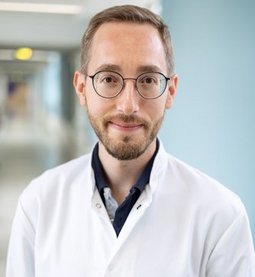 Dominik Zach (© Johannes Lehner) Portrait eines schmalen Herren mit hohem braunen Haaransatz, kurzem Vollbart, runder Brille, lächelnd