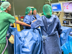 Tag der Ausbildung Gummibärchen-OP (© Sandra Wimmer) Drei Mädchen in OP-Kleidung- blaue Kitteln und grünen Hauben vor dem OP-Tisch mit den beiden Monitoren auf dem die Gummibärchen im inneren der Puppe zu sehen sind. Ein Pfleger gibt Anweisungen am Kopf der Puppe stehend.