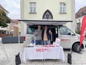 Weißer Hörbus vor Turm mit Markise und Tisch mit weißer Tischdecke davor, darauf gedruckt "MED-EL" mit Flyern und Give-Aways. Links in grauem Pulli und Hemd, rechts mit blauer Steppweste, beigem Pulli und Brille, beide in Jeans