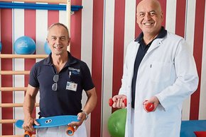 Klinik Geriatrie (© Monika Schneider-Stranninger) Der kleinere Herr links mit dunkelblauem Polohemd und weißer Hose hält ein kleines blaues Skateboard, der größere kräftiger Herr rechts mit Glatze zwei kleine rote Hanteln. beide vor rot-weiß gestreifter Tapete mit einem Regal mit Sportutensilien im Hintergrund, in die Kamera lachend.