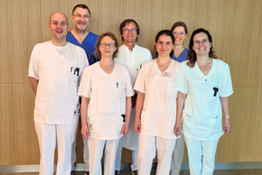 Die Gruppe vor einem Holzpaneel alle in weiß, Prof. Weigert und Frau Bischof in blau