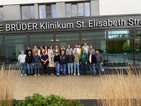 Erster pPOCUS KUrs Gruppenbild (© Dr. Thomas Irlbeck) Vor dem Haupteingang in zwei Reihen, im Vordergrund beige Gräser links und rechts, mittig etwas Grün. Viele In Jeans.