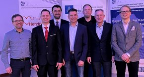 Im leicht lila Beamerlicht bis auf den Herrn links alle in Sacko, lachend, vor Rollups