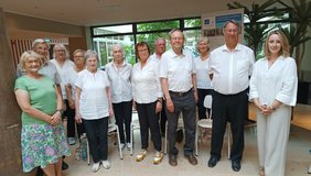 Bachsingers (© Rita Roth) Alle Sänger und Sängerinnen fortgeschrittenen Alters mit weißem Oberteil und dunkler Hose. Frau Urban links in grün mit Rock und Frau Korber rechts mit beiger Hose. alle im Foyer mit grünem Hintergrund- Sicht zum Innenhof.