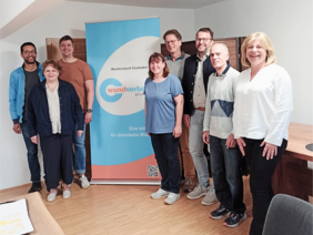 Das Team auf einem Holzboden mit schräger Decke neben einem blau-roten Rollup stehen