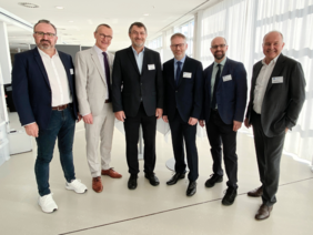 Leiter der Krebszentren (© Sandra Wimmer) Bis auf einen sind alle Herren in dunklem Anzug mit dunkler Hose oder Jeans. Der zweite von links ist mit beigem Anzug und braunen Schuhen zu sehen. Vier davon tragen Brille. Der Hintergrund ist eine große Fensterfront mit Vorhan g und hellgbeigem Boden und Decke, modern. Alle schauen freundlich drein.