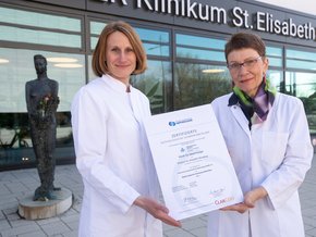 Zertifizierung Schwerpunktklinik Nephrologie (© Johannes Lehner) Zwei Damen im Kittel, links mit halblangem Haar, rechts mit kurzem Haar, Brille und Halstuch. Beide halten ein Zertifikat in DINA3 und lächeln. Außen vor dem Klinikumseingang stehend.