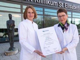 Zwei Damen im Kittel, links mit halblangem Haar, rechts mit kurzem Haar, Brille und Halstuch. Beide halten ein Zertifikat in DINA3 und lächeln. Außen vor dem Klinikumseingang stehend.