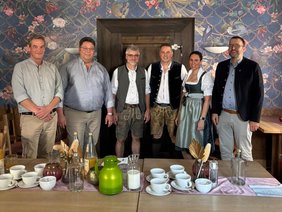 Trauma-Fortbildung (© Klinikum Straubing) Foto im Wirtshaus vor einem dekorativ gedeckten Tisch mit bunter Tapete und rustikaler Tür im Hintergrund. Bis auf den Herrn rechts und den beiden links sind die Teilnehmer in Tracht.