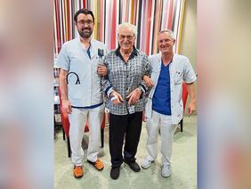 Den Tod überlebt (© Thomas Heigl) Weißhaariger Herr mit Brille und Satinpyjama in grau/schwarz mit zwei Ärzten am Arm, alle lachen. Im Hintergrund bunt gestreifter Vorhang