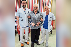 Weißhaariger Herr mit Brille und Satinpyjama in grau/schwarz mit zwei Ärzten am Arm, alle lachen. Im Hintergrund bunt gestreifter Vorhang