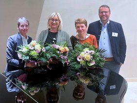 Alle vor einem Klavier, die Damen links mit großen Blumensträußen, zwei mit kurzen Haaren, alle lächelnd.
