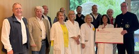 Spende Lionsclub Straubing Gelbe Damen und Herren (© Dr. Christian Waas) Gruppenfoto im Foyer vor günem LED Bild, viele weiß oder dunkel gekleidet. Die Dame rechts vorne zeigt einen großen Spendenscheck.