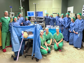 Tag der Ausbildung OP (© Sandra Wimmer) Schüler in blauen OP-Kitteln und grüner Haube hinter dem OP-Tisch stehend, links der Pfleger und vorne die drei Pflegerinnen jeweils in grünen Kasaks und Hosen.