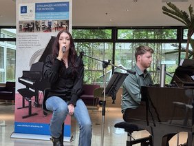 Auf dem Bild sind zwei Musikerinnen und Musiker zu sehen: Rika Mages und Michi Stieglmeier. Rika Mages sitzt vorn und singt. Michi Stieglmeier begleitet sie mit einem Klavier. Beide treten gemeinsam als Duo SoulSync auf. Sie befinden sich bei einem Konzert im Rahmen von „Straubinger Musizieren für Patienten“. Im Hintergrund ist das Foyer des Klinikums  zu sehen.