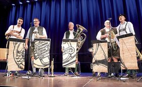 Auf der Bühne vor dunkelblauem Vorhang Männer in Lederhose, weißes Hemd, zum Teil Westen und Trompeten und Posaunen singend
