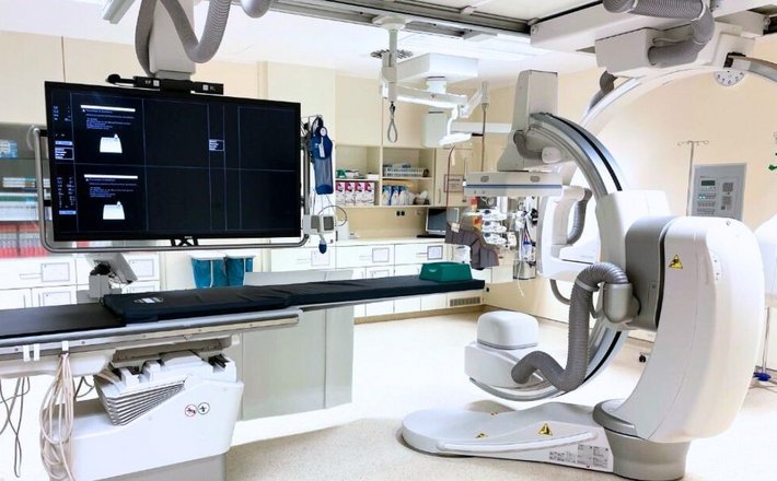 Moderne Angiografie-Anlage in einem klinischen Untersuchungsraum. Die Anlage besteht aus einem großen C-Bogen mit integriertem Flachdetektor, einem höhenverstellbaren Untersuchungstisch und mehreren Monitoren in einem großen Monitor zur Bilddarstellung. Der Raum ist hell und technisch hochwertig ausgestattet, mit Deckenarmen für Kabel und zusätzlichen medizinischen Geräten.