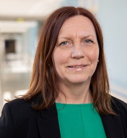 Andrea Gerstl (© Johannes Lehner) Dame mit braunen mittellangen Haaren freundlich lächelnd mit dunkelgrünem Oberteil und dunklem Jackett vor hellblau-weißem Klinikflur