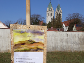 Ein Schild an einem Holzblock mit Bild eines Kreuzes und Morgenhimmel sowie Text. Im Hintergrund sieht man die zwei Türme der St. Peterskriche