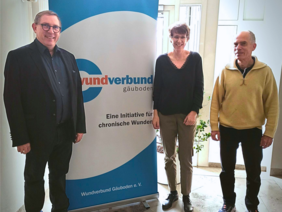 Rezertifizierung Wundexperten (© Sabine Muck) Links ein Mann in dunklem Anzug und Brille, rechts ein Herr in gelben Pulli und mittig die Dame mit kurzem Haar und V-Ausschnittpulli vor dem Wunderverbund-Rollup. Alle lächelnd.