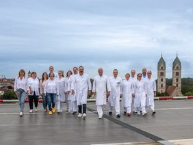 Team der Klinik für Allgemein-, Viszeral- und Minimalinvasive Chirurgie und Proktologie (© Johannes Lehner) Viele in Arztkittel, links die Damen zum Teil zivil in Jeans, ca . die Hälfte Frauen. Auf dem Hubschrauberlandeplatz bei bewölktem Himmel mit den zwei St. Peterskirchen-Türmen im Hintegrund, alle auf den Fotografen zugehend