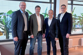 Vor einer Glasfront im Foyer mit Pflanzen im Hintergrund, die beiden Herren außen größer und mit Anzug, links mit Glatze, Vollbart, Brille, rechts mit kurzen braunen Haaren, Brille, alle Sakkos, freundlich lächelnd