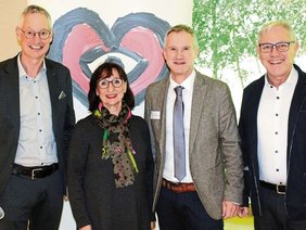 Vor dem RollUp der Herzstiftung die Herren mit Jackett, rechts mit Jacke, Frau Mehr am kleinsten mit schwarzem Pulli und buntem Schal. Alle Brille und lachend.