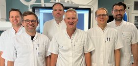Alle Ärzte im Herzkatherraum von dem Bildschirm des Geräts mit kurzärmeligen weißen Kitteln, lächelnd