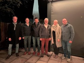 Alle in Jacke, zum Teil Schal, viele in Jeans, vor dunklem Hintergrund. Die zwei Herren links und der rechts mit Brille