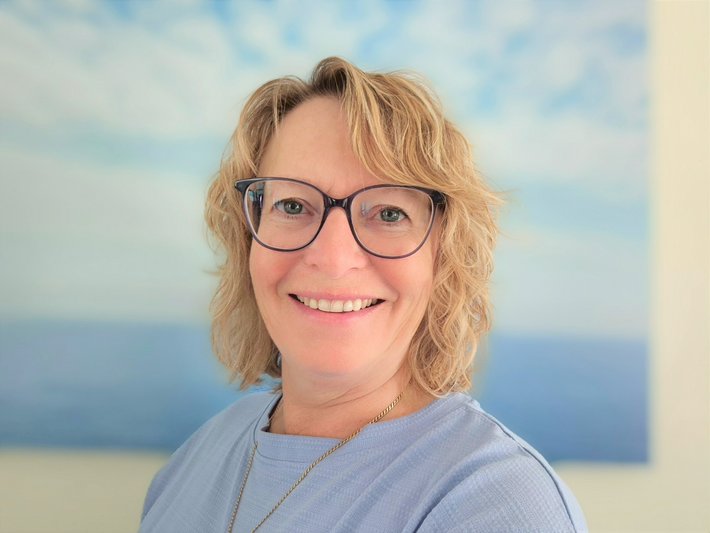 Daniela Müller, (© Klinikum Straubing) Blonde Frau mit halblangen Locken und großer Cateye-Brille mit dunklem Rahmen, Kette, blauer Pulli vor einen Bild mit Strand und blau-weißem Himmel