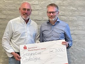 Leukämiehilfe Ostbayern Spende (© Prof. Markus Völk) Zwei Herren in Jeans und Hemd mit großem Spendenscheck vor einer Hauswand