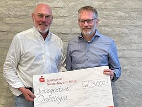 Leukämiehilfe Ostbayern Spende (© Prof. Markus Völk) Zwei Herren in Jeans und Hemd mit großem Spendenscheck vor einer Hauswand