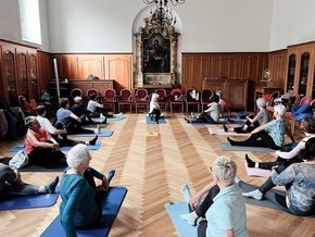 Kloster Azlburg Integrative Onkologie (© Stephanie Seckel) Ein langer Raum mit kleinem Wandaltar in der Mitte und Kronleuchter, halb holzvertäfelte Wände und Parkett mit Yogateilnehmenden auf Matten am Boden sitzen, stretchend.