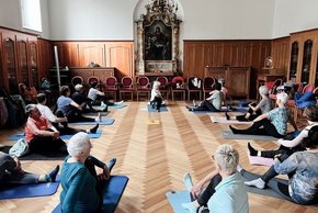Kloster Azlburg Integrative Onkologie (© Stephanie Seckel) Ein langer Raum mit kleinem Wandaltar in der Mitte und Kronleuchter, halb holzvertäfelte Wände und Parkett mit Yogateilnehmenden auf Matten am Boden sitzen, stretchend.