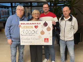 Die blonde Dame mit schwarzer runder Brille und großem Scheck mit Überschrift "SOLIDARITÄT LEBEN" schwarzer weiter Hose, Sneakers, die Herren in Pulli und Jeans